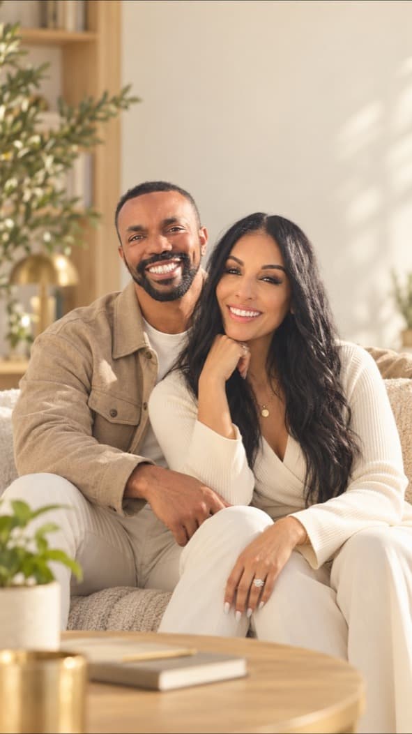Alfonso and Vanessa Garcia sitting together on a cream sofa in warm daylight, smiling at the camera